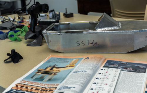 Small welded metal boat sits on a school desk with an instructional textbook and mechanical parts around it.