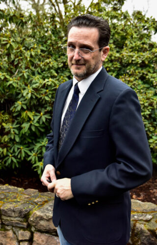 Man standing in front of a rock bench wearing glasses dressed in a suit.