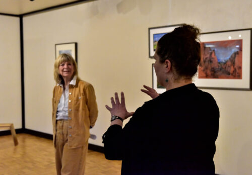 A woman stands farther back with her arms clasped calmly behind her with an smile. In front of her facing to the right is a woman whose hands are in front of her. They are in the art studio with two pictures mounted on the wall on the left of them.