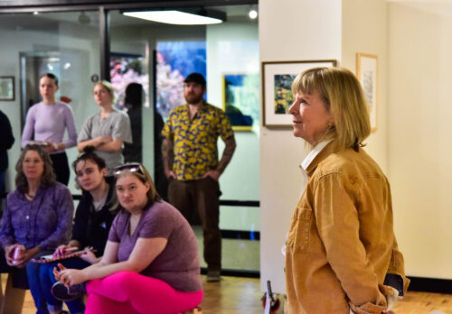 Woman stands in the UCC art studio. She is  facing the left wearing a light brown jacket. Her arms are  casually clasped behind her. A small group, with some standing and some sitting, face forward. 