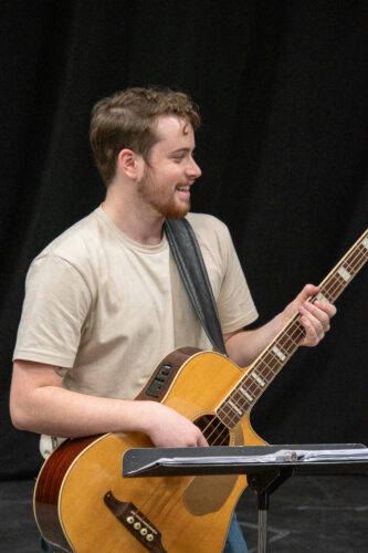 Guy with a slight beard and a smile plays on his guitar. He is standing in front of a black curtain with a music stand in front of him.