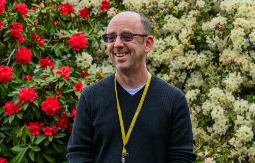 Man wearing dark glasses smiles to the right. Behind him on the right are red flowers with white flowers on the left.. He is wearing a long navy blue shirt with a light blue shirt peeking out at his collar. There is a yellow lanyard around his neck.