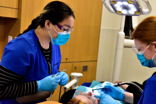 Two dental students stand on either side of person who is laying down. The students are wearing medical shirts and mouth coverings. They are in the process of cleaning teeth.