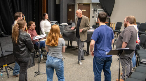 Inside the Whipple Art Center, students form a semi-circle around a professor. He is standing facing towards the right talking to a student. He is wearing a tan suit jacket, black shirt,  grey jeans and tennis shoes. He is standing in front of a piano and music stands are placed near students. To the left of the students facing their back is a row of chairs.