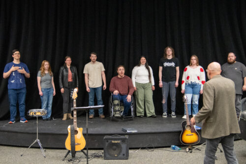 On the Whipple Fine Art's stage, all students (besides one) stand to sing. The professor, wearing a tan jacket and grey slacks, is conducting. To either side of the stage are guitars and one speaker.