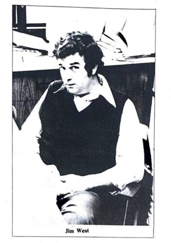 Man casually sits down with his hands on his lap. He is wearing a long sleeve buttoned up shirt and a vest. He looks up at the camera with a unsure glance.