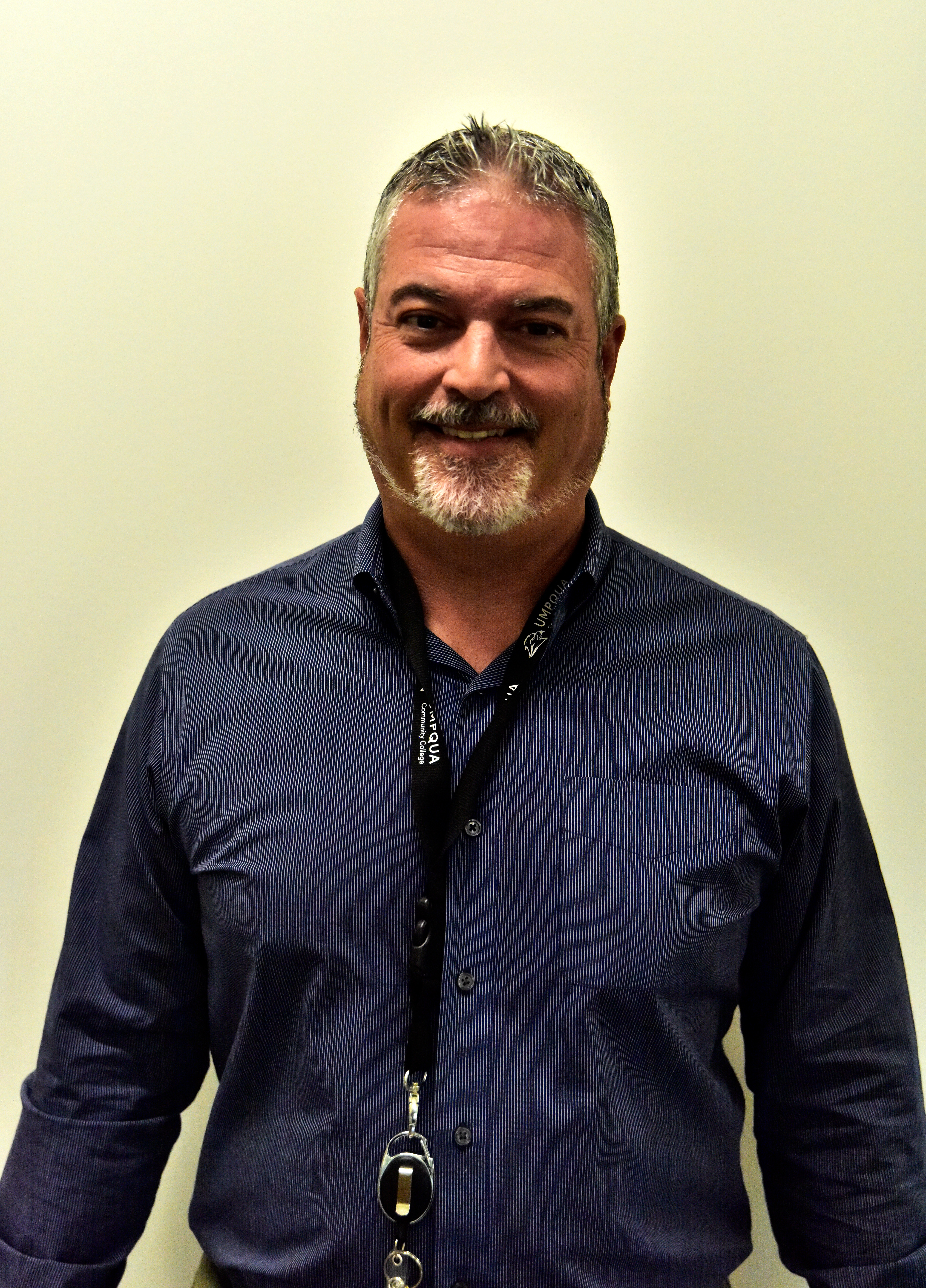 A man is standing with a smile in front of a plain off-white wall. He has short grey and white hair and a goatee that is predominantly white. He is wearing a long sleeved blue shirt with a black lanyard around his neck.
