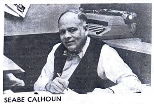 Older man sits down at his desk. He is smiling and wearing a dark vest and stripped bowtie. An old fashioned typewriter is on a shelf behind him.