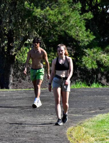 Two people walk on a curved track towards the camera. One person is wearing shorts and white tennis shoes. The second person is wearing a sports bra, shorts, and black tennis shoes. Trees create shadows for them in the background.
