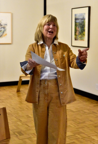 Woman stands in an art studio with a painting on either side of the wall behind her. She is holding a paper in her hand and pointing into the audience as she speaks. She is wearing a white shirt, a light brown jacket and matching pants.