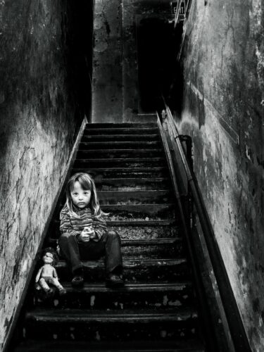 Little girl sits on a staircase with an old doll. She is wearing a long-sleeved shirt and pants. The staircase goes upward and two walls stand on either side of it.  It is a black and white image.