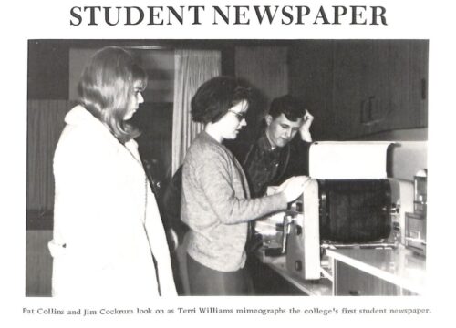 Two students stand, one rests his head against his hand, and watch as another student mimeographs the student newspaper. They are in casual attire. 