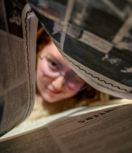 A page of newspaper is folded into a tent shape. A student peers through these pages facing the camera. He is wearing prescription gasses and has red hair.